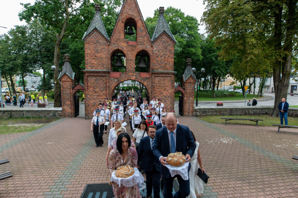 Grupa ludzi w eleganckich strojach niesie ozdobny chleb, idąc przez ceglaną bramę. W tle zielone drzewa i uczestnicy wydarzenia. Sceneria jest na zewnątrz, na wybrukowanym placu.