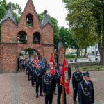 Na zdjęciu widoczny jest pochód strażaków w czarnych mundurach z czerwonymi sztandarami. W tle znajduje się ceglany budynek z trzema wieżyczkami i dzwonami. Otoczenie to zewnętrzna przestrzeń z drzewami.