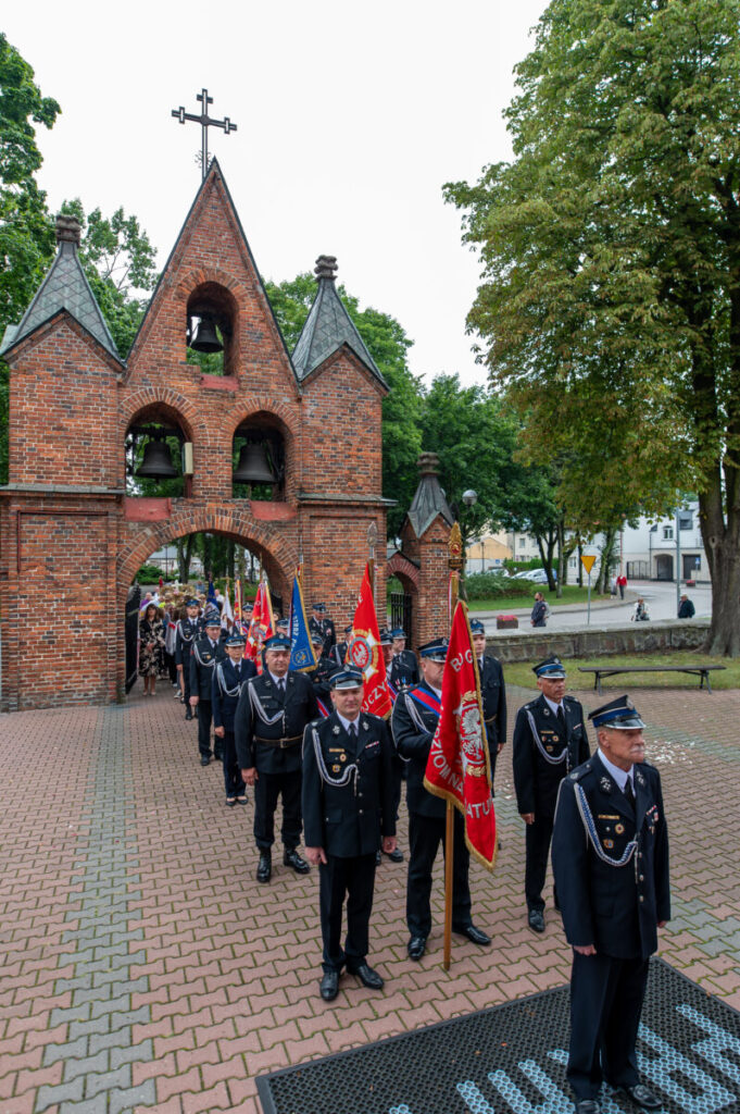 Na zdjęciu widoczny jest pochód strażaków w czarnych mundurach z czerwonymi sztandarami. W tle znajduje się ceglany budynek z trzema wieżyczkami i dzwonami. Otoczenie to zewnętrzna przestrzeń z drzewami.