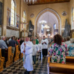 Procesja w kościele katolickim; białe szaty, kolorowe witraże, złote żyrandole; ludzie stoją w ławkach po bokach.