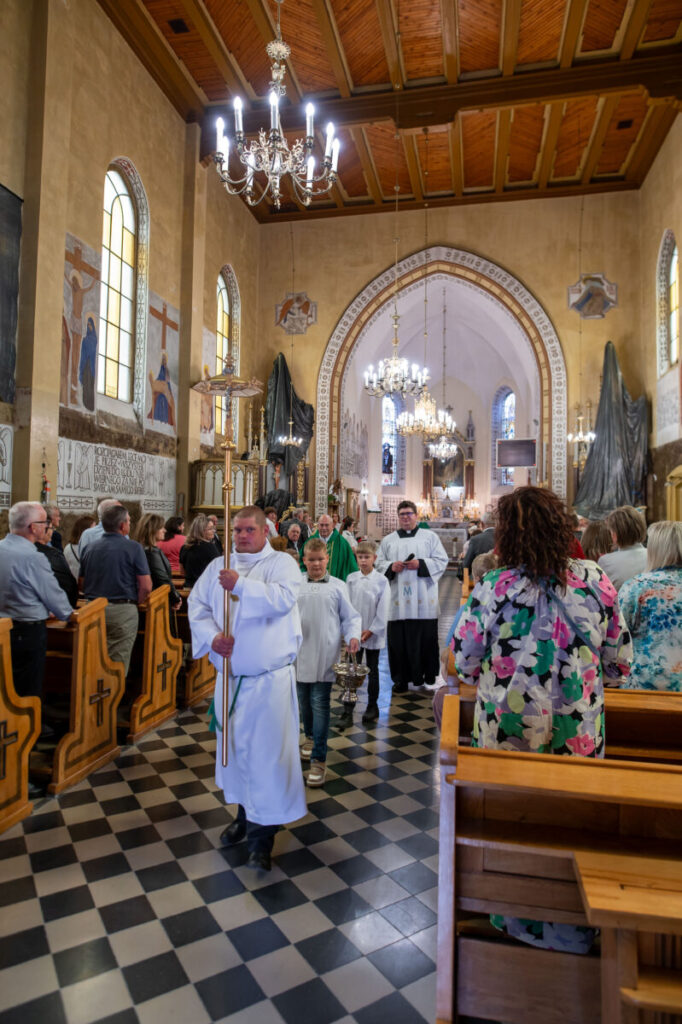 Procesja w kościele katolickim; białe szaty, kolorowe witraże, złote żyrandole; ludzie stoją w ławkach po bokach.