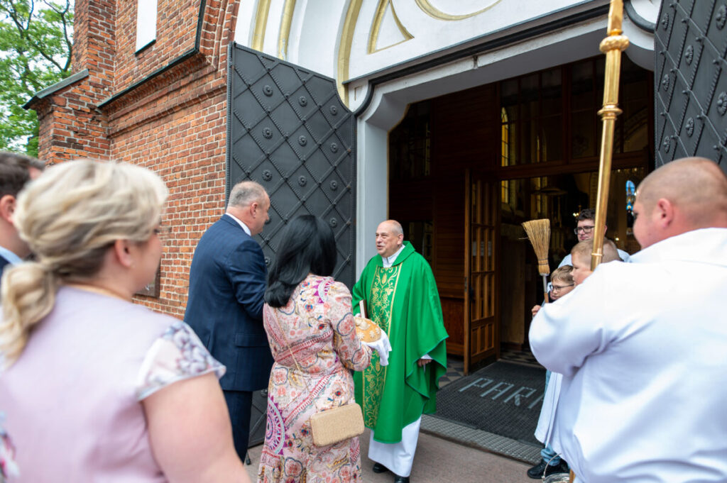 Osoba w zielonej szacie liturgicznej stoi przed kościołem z cegły. Wokół niej kilku ludzi, w tym dzieci. Widoczne są detale architektoniczne drzwi.