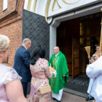 Osoba w zielonej szacie liturgicznej stoi przed kościołem z cegły. Wokół niej kilku ludzi, w tym dzieci. Widoczne są detale architektoniczne drzwi.