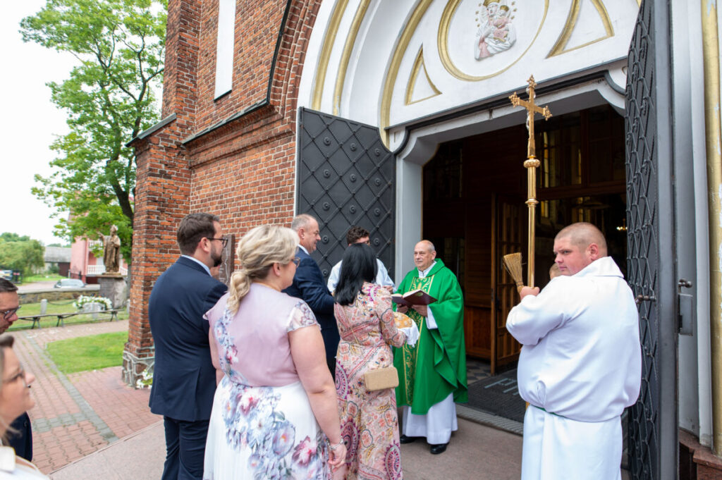 Grupa ludzi przed kościołem rozmawia z księdzem w zielonej szacie. Budynek z cegły. Widać dekoracje i drzewa.
