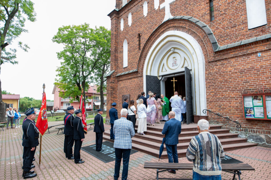 Kościół z czerwonej cegły, ludzie wchodzą przez drzwi, wokół drzewa. Grupa osób w strojach galowych trzyma czerwone flagi.