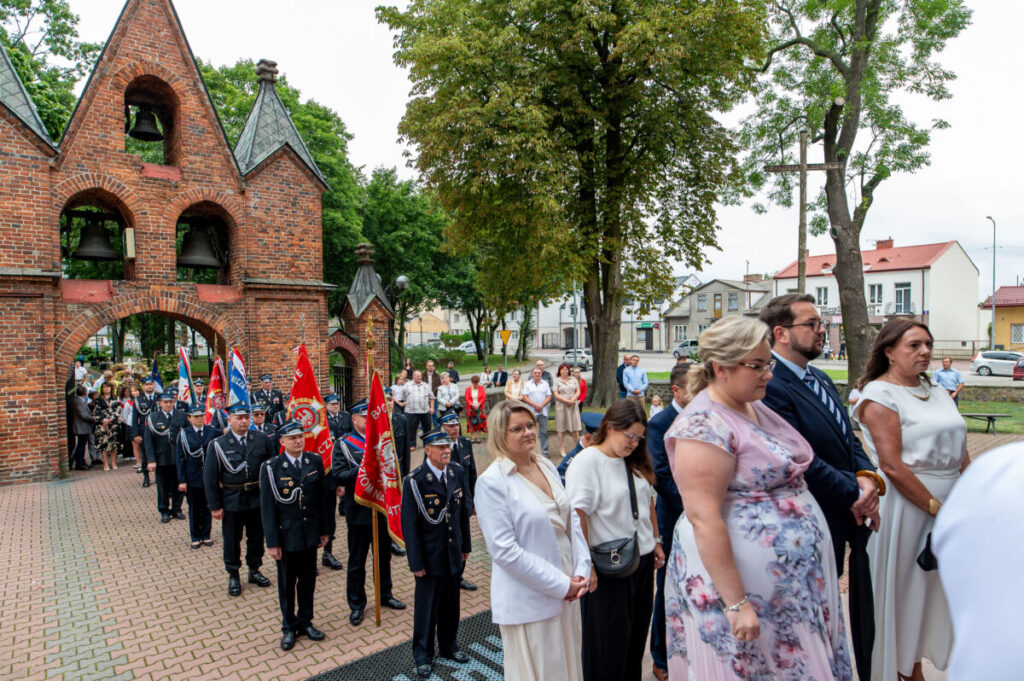 Grupa ludzi w eleganckich ubraniach stoi przed ceglaną bramą z dzwonami. Widać czerwone i niebieskie flagi. W tle drzewa oraz budynki na ulicy.