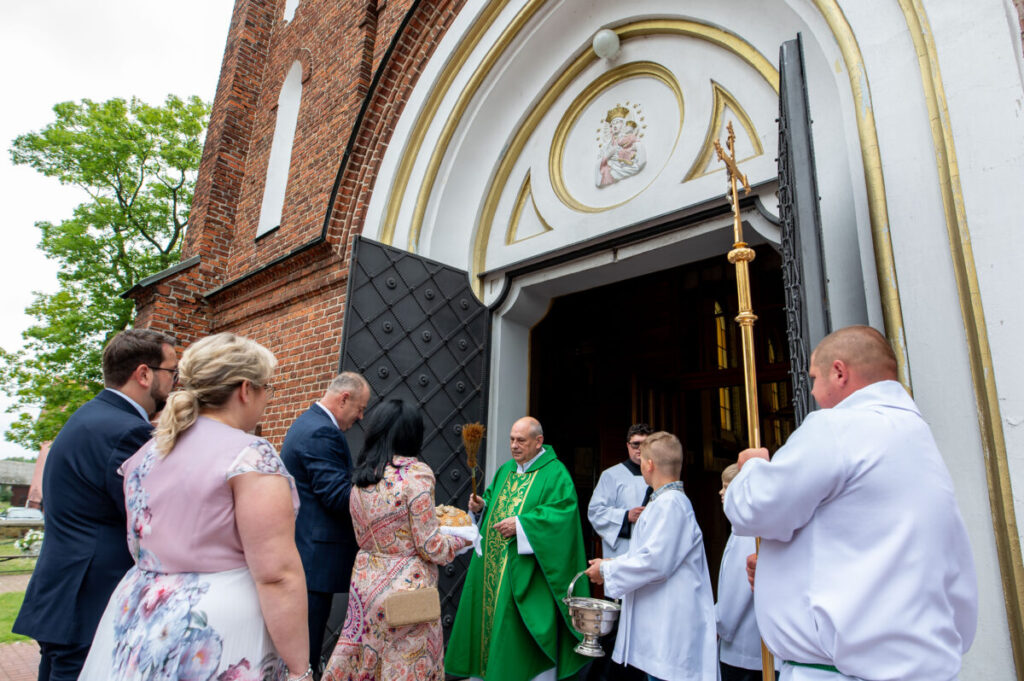 Grupa ludzi w strojach liturgicznych przed kościołem z cegły. Kapłan w zielonej szacie błogosławi uczestników. Tło zdobią drzwi z ornamentami i medalion z wizerunkiem Madonny.