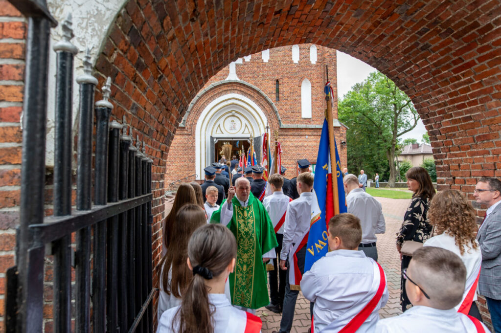 Grupa ludzi w białych koszulach z flagami i ksiądz w zielonym ornacie przed kościołem z cegły. Tło zielone drzewa. Scena ceremonii religijnej.