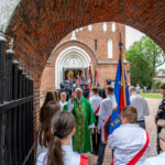 Grupa ludzi w białych koszulach z flagami i ksiądz w zielonym ornacie przed kościołem z cegły. Tło zielone drzewa. Scena ceremonii religijnej.