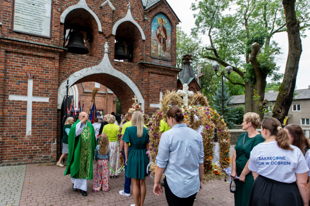 Na zdjęciu widoczny jest ksiądz w zielonym ornacie przed murowanym kościołem z dzwonami. Wokół niego znajdują się ludzie, niektórzy w odświętnych strojach, trzymający wieńce dożynkowe. Tło stanowią