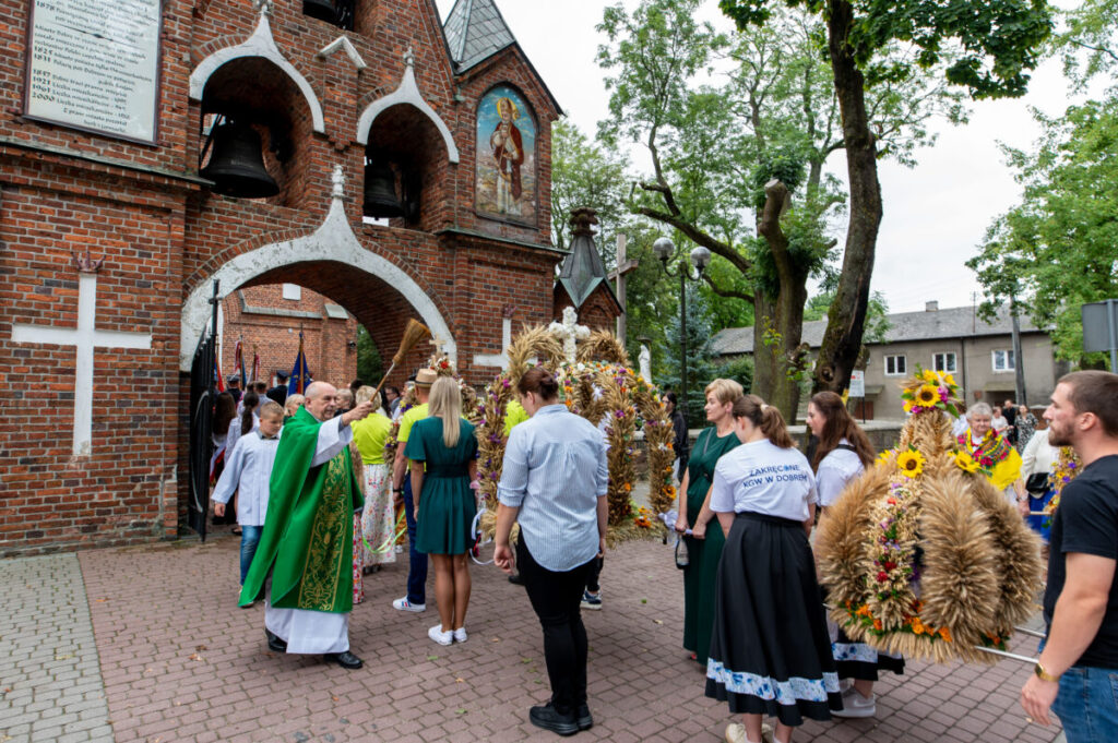 Procesja przed ceglaną świątynią; kapłan błogosławi uczestników w kolorowych ubraniach. Otoczenie: zielone drzewa.
