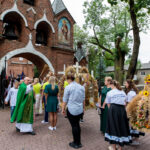 Procesja przed ceglaną świątynią; kapłan błogosławi uczestników w kolorowych ubraniach. Otoczenie: zielone drzewa.