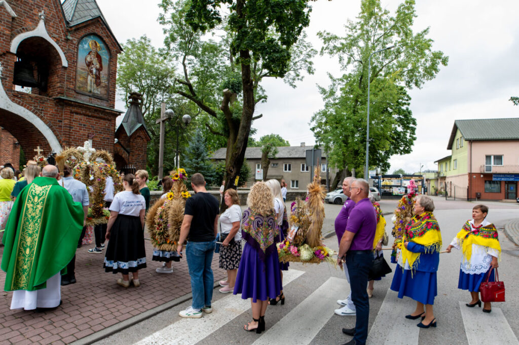 Grupa ludzi z wieńcami dożynkowymi stoi przed zabytkowym kościołem z czerwonej cegły. Ubrani są w kolorowe stroje ludowe. Tło zdobią zielone drzewa i budynki.