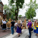 Grupa ludzi z wieńcami dożynkowymi stoi przed zabytkowym kościołem z czerwonej cegły. Ubrani są w kolorowe stroje ludowe. Tło zdobią zielone drzewa i budynki.