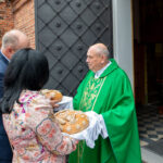 Mężczyzna w zielonej szacie przyjmuje tradycyjny chleb od dwojga ludzi. Tło to ceglany budynek i czarne drzwi. Sytuacja odbywa się na zewnątrz.