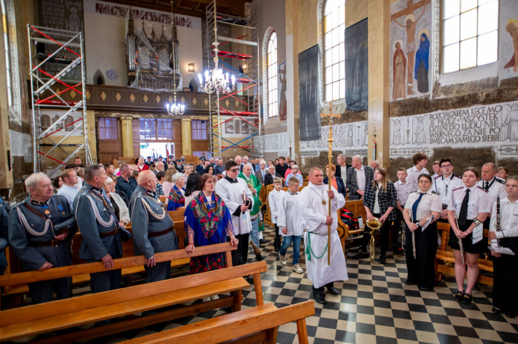 Grupa ludzi w kościele, ubrani formalnie, uczestniczą w ceremonii. W tle widać malowidła, okna i rusztowanie. Na podłodze wzory w szachownicę.