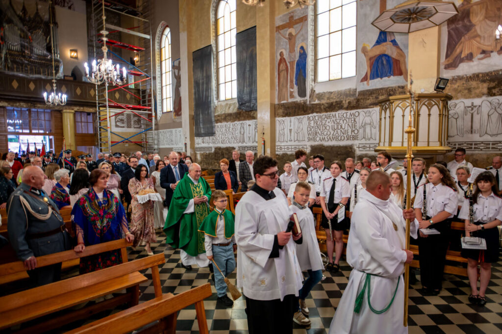 Procesja w kościele, ludzie w białych i zielonych szatach. Tło z freskami i żyrandolami. Uczestnicy stoją na czarno-białej posadzce.
