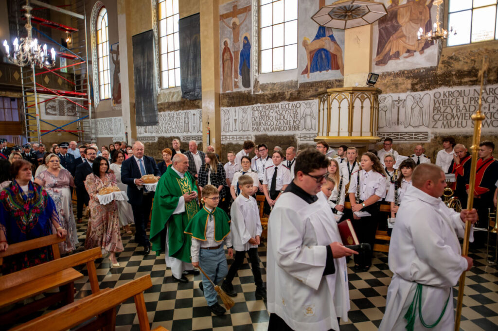 Odprawa kościelna z udziałem wiernych. Ludzie w kolorowych i białych strojach na tle fresków i witraży. Uroczysta atmosfera.