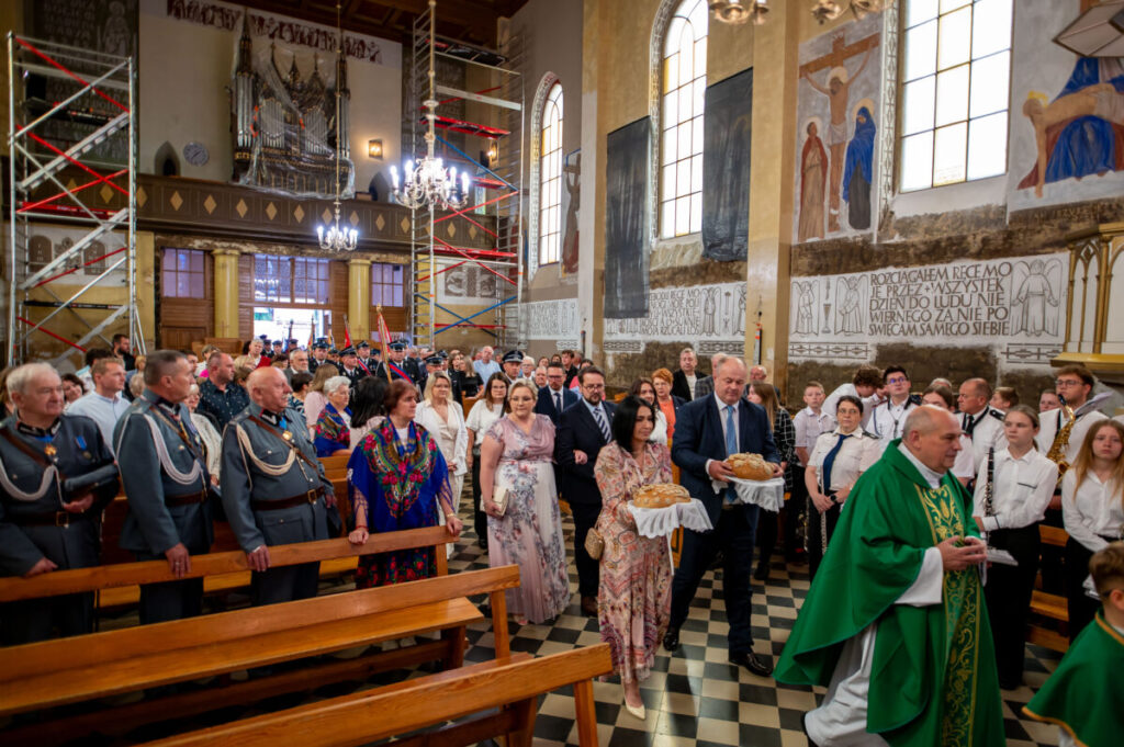 Ludzie w odświętnych strojach niosą chleb w kościele. Ściany ozdobione malowidłami i napisami, tło w ciepłych kolorach. Zdjęcie pełne detali, pokazuje uroczystość religijną.
