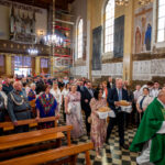 Ludzie w odświętnych strojach niosą chleb w kościele. Ściany ozdobione malowidłami i napisami, tło w ciepłych kolorach. Zdjęcie pełne detali, pokazuje uroczystość religijną.