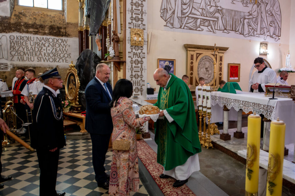 Na zdjęciu ksiądz w zielonym ornacie przyjmuje chleb od pary w kościele. Wnętrze jest ozdobione freskami i wzorami w ciepłych kolorach. W tle widoczne są świece i instrumenty dęte.