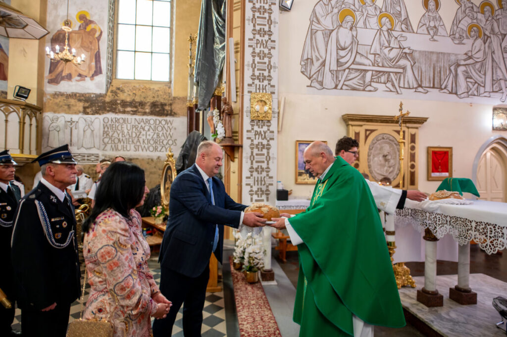 Ceremonia w kościele, mężczyzna w garniturze przekazuje chleb księdzu w zielonej szacie. W tle malowidła i ozdoby. Obecni strażacy w mundurach.