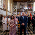 Ludzie stoją w kościele, kobieta w kolorowej sukni, mężczyzna w garniturze. W tle sklepione okna i sztandary. Na ścianach freski.