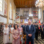 Grupa ludzi w eleganckich strojach stoi w kościele z drewnianym sufitem. Podłoga ma czarno-białe kafelki. W tle widoczne są barwne witraże i żyrandole.