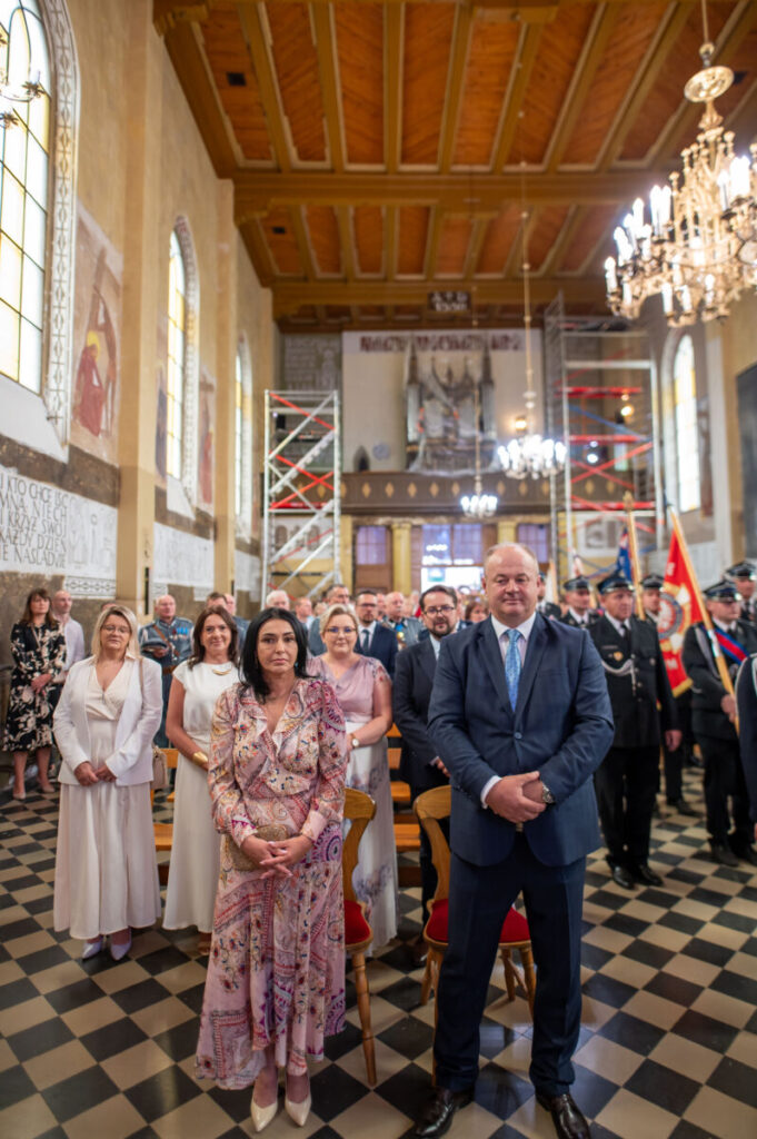 Grupa ludzi w eleganckich strojach stoi w kościele z drewnianym sufitem. Podłoga ma czarno-białe kafelki. W tle widoczne są barwne witraże i żyrandole.