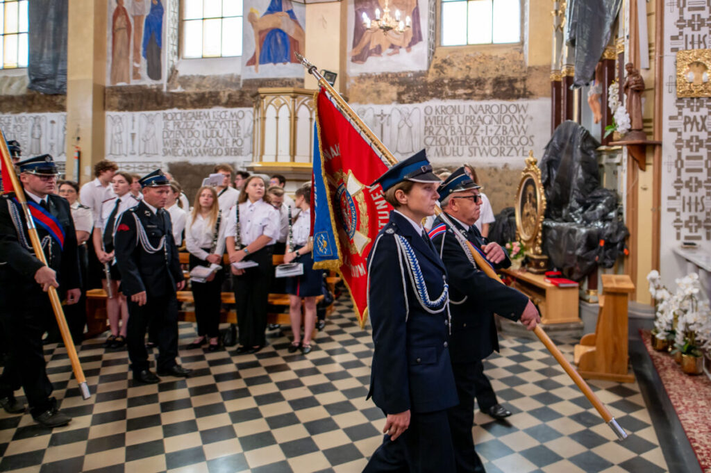 Na zdjęciu widoczni są strażacy w mundurach z czerwonym sztandarem w kościele. Posadzka czarno-biała, ściany zdobione freskami. W tle stoi grupa osób w białych koszulach.