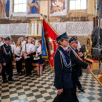 Na zdjęciu widoczni są strażacy w mundurach z czerwonym sztandarem w kościele. Posadzka czarno-biała, ściany zdobione freskami. W tle stoi grupa osób w białych koszulach.