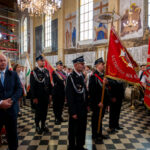 Wnętrze kościoła, grupa strażaków w mundurach z czerwonymi sztandarami. Tło zdobią kolorowe freski i żyrandole. Na podłodze czarno-biała szachownica.