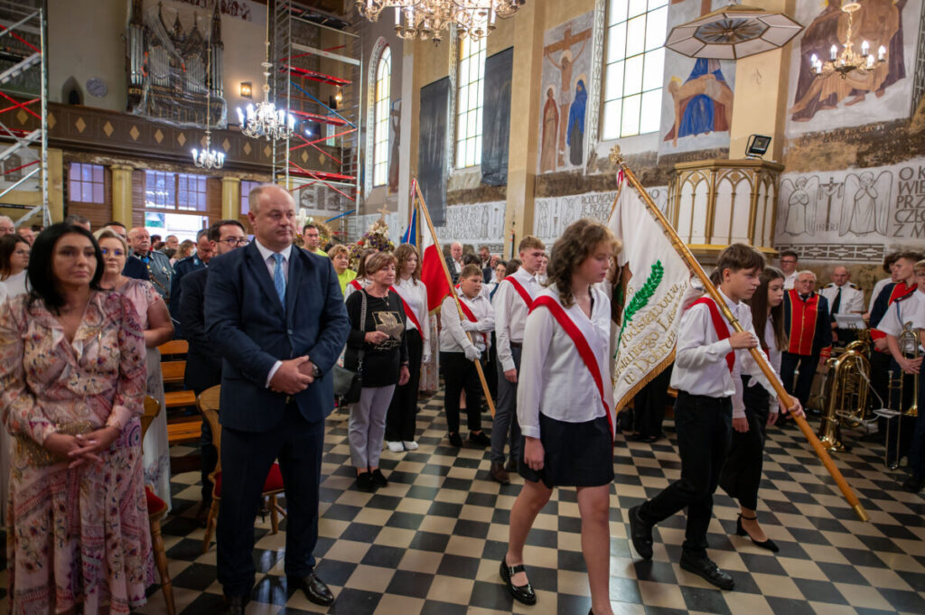 Grupa ludzi w kościele, młodzież z sztandarami, eleganckie ubrania. Wnętrze zdobione freskami i żyrandolami. Atmosfera uroczysta.
