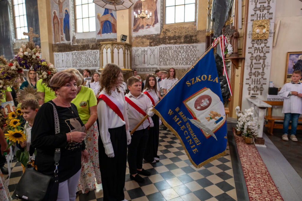 Grupa osób stoi w kościele, trzymając niebieski sztandar szkoły. Wnętrze zdobią freski i drewniane elementy. Tło jest jasne z dekoracjami.