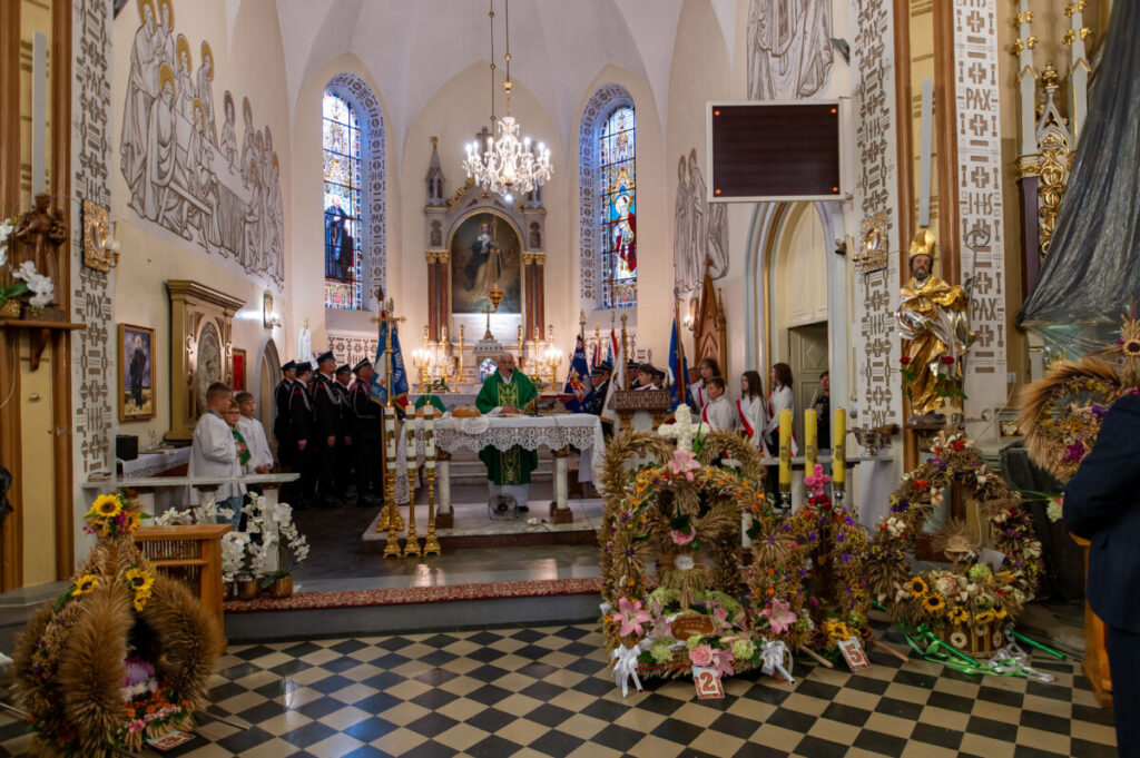 Wnętrze kościoła z uroczystością. Na pierwszym planie wieńce dożynkowe, w tle ołtarz z witrażami i malowidłami, dominują jasne kolory. Po prawej osoby w białych szatach liturgicznych.