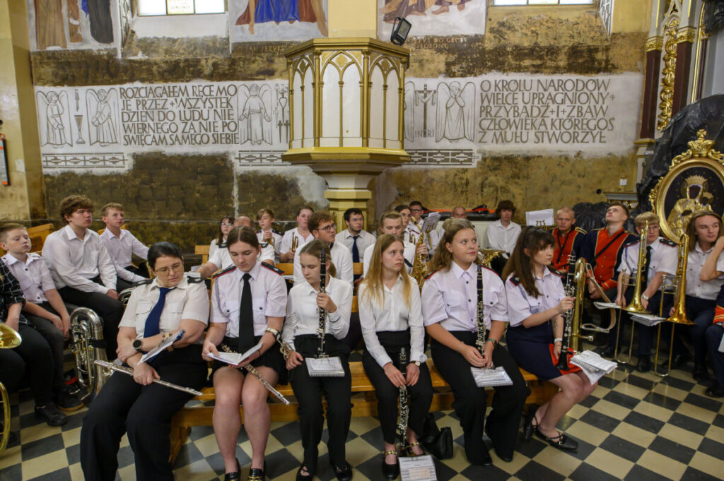 Grupa młodych muzyków w białych i ciemnych strojach siedzi w zabytkowym wnętrzu. Ściany ozdobione są freskami i napisami. Scena mieści się na tle kamiennej architektury.