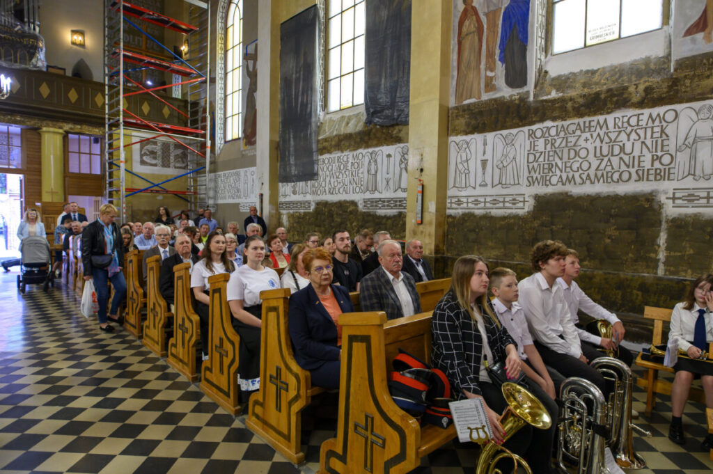 Grupa ludzi siedzi w ławkach kościoła z malowidłami na ścianach. Na pierwszym planie widać młodzież z instrumentami muzycznymi. W tle jasne ściany, okna i są rusztowania.