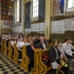 Grupa ludzi siedzi w ławkach kościoła z malowidłami na ścianach. Na pierwszym planie widać młodzież z instrumentami muzycznymi. W tle jasne ściany, okna i są rusztowania.