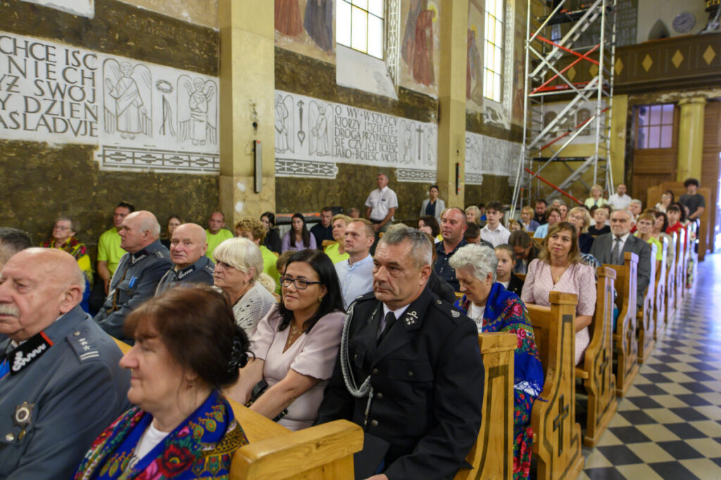 Na zdjęciu widzimy ludzi siedzących w kościelnych ławkach. Ściany ozdobione są freskami i tekstami w jasnych tonach. W tle widać rusztowanie, co sugeruje remont.