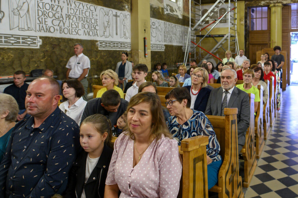 Grupa ludzi siedzi w ławkach w kościele. W tle widać dekoracje ścienne z napisami. Tło jest w kolorach brązu i beżu.