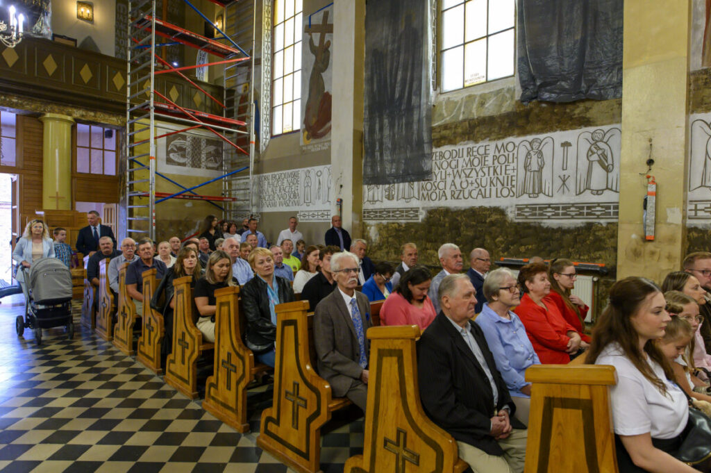 Kościół pełen ludzi siedzących w drewnianych ławkach, na tle jasnych ścian z malowidłami i napisami. Widać rusztowanie oraz okna wpuszczające światło. Podłoga jest w czarno-białe wzory szachownicy.