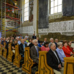 Kościół pełen ludzi siedzących w drewnianych ławkach, na tle jasnych ścian z malowidłami i napisami. Widać rusztowanie oraz okna wpuszczające światło. Podłoga jest w czarno-białe wzory szachownicy.