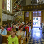 Grupa ludzi siedzi na ławkach w kościele z freskami na ścianach. W tle widać otwarte drzwi i osoby na zewnątrz. Wnętrze zdobią kolorowe witraże i dekoracje.
