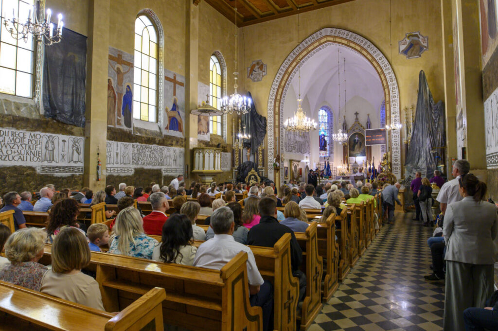 Wnętrze kościoła z wiernymi na ławkach, jasne witraże i freski na ścianach. Żyrandole oświetlają pomieszczenie. Msza święta trwa.