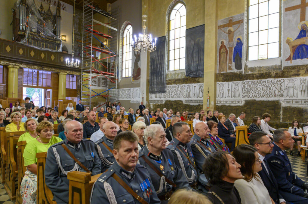 Grupa ludzi siedzi w kościele z freskami na ścianach. W tle widać rusztowanie i oświetlenie z żyrandola. Wnętrze jest jasne, dominują kolory beżowy i niebieski.