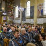 Grupa ludzi siedzi w kościele z freskami na ścianach. W tle widać rusztowanie i oświetlenie z żyrandola. Wnętrze jest jasne, dominują kolory beżowy i niebieski.