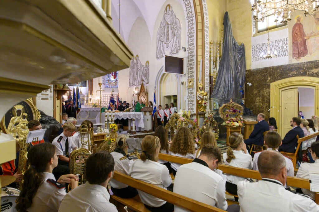Grupa ludzi siedzi w kościele z jasnym wystrojem wnętrza. Na ołtarzu jest kapłan w zielonej szacie. Po lewej stronie widać orkiestrę z instrumentami dętymi.