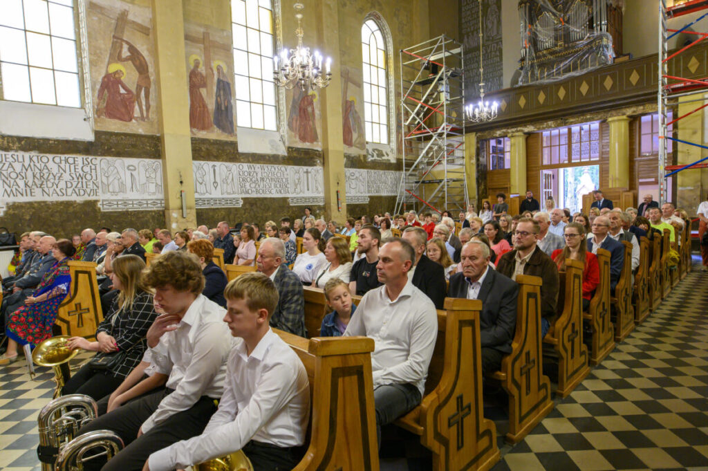 Grupa osób siedzi w kościele z drewnianymi ławkami, muralami i organami w tle. Dominują kolory brązowy i złoty. Na ścianach widoczne są freski i teksty.
