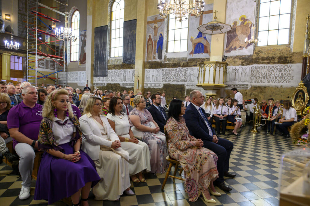 Grupa ludzi siedzi w kościele. Ściany zdobione malowidłami i napisami. Dominują kolory beżowy i różne odcienie fioletu.