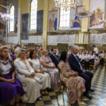 Grupa ludzi siedzi w kościele. Ściany zdobione malowidłami i napisami. Dominują kolory beżowy i różne odcienie fioletu.